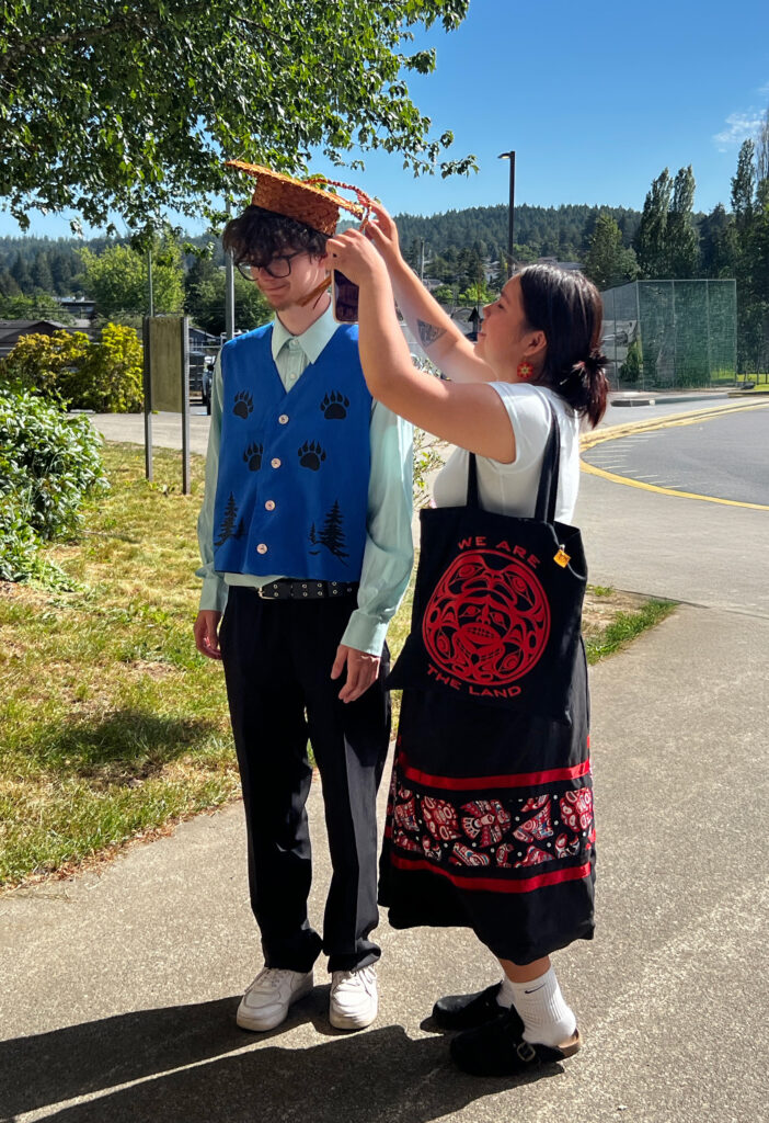 Young woman in a ribbon skirt helps a young man with his wovengrad cap