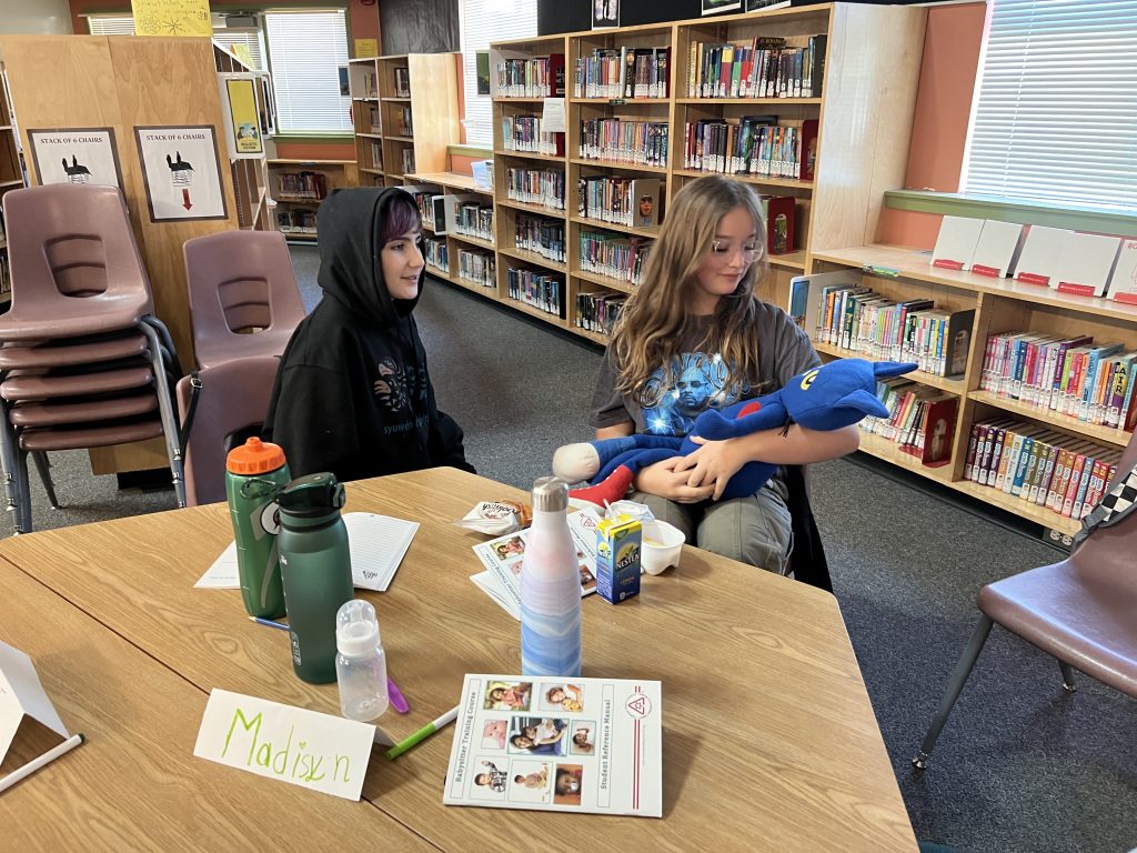 Two middle-school aged kids sit in the library, one is cradling a stuffed cat as if it's a baby to show what she's learned in the babysitter's course