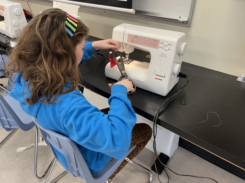 A girl seated at a sewing machine holds scissors to cut a piece of thread. Phot taken over her shoulder from behind and to the side so her face is not shown